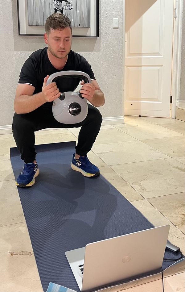 Someone doing a kettlebell squat at home, with their coach live on a laptop on the floor in front of them